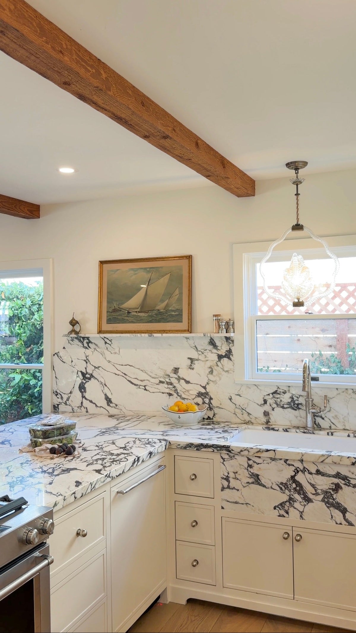 A kitchen interior with white cabinets, white marble countertops and backsplash with black veining, a hanging pendant light, a framed sailboat painting on the wall, and exposed wooden ceiling beams.